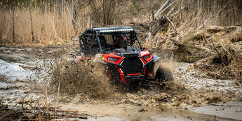 Polaris RZR 1000 XP UTV featuring two people in action navigating a large mud hole. The image showcases the durability of the impact-resistant Perfex products.